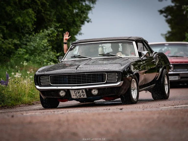 Svart Chevrolet Camaro 1969 passerar fotografen och passageraren vinkar.